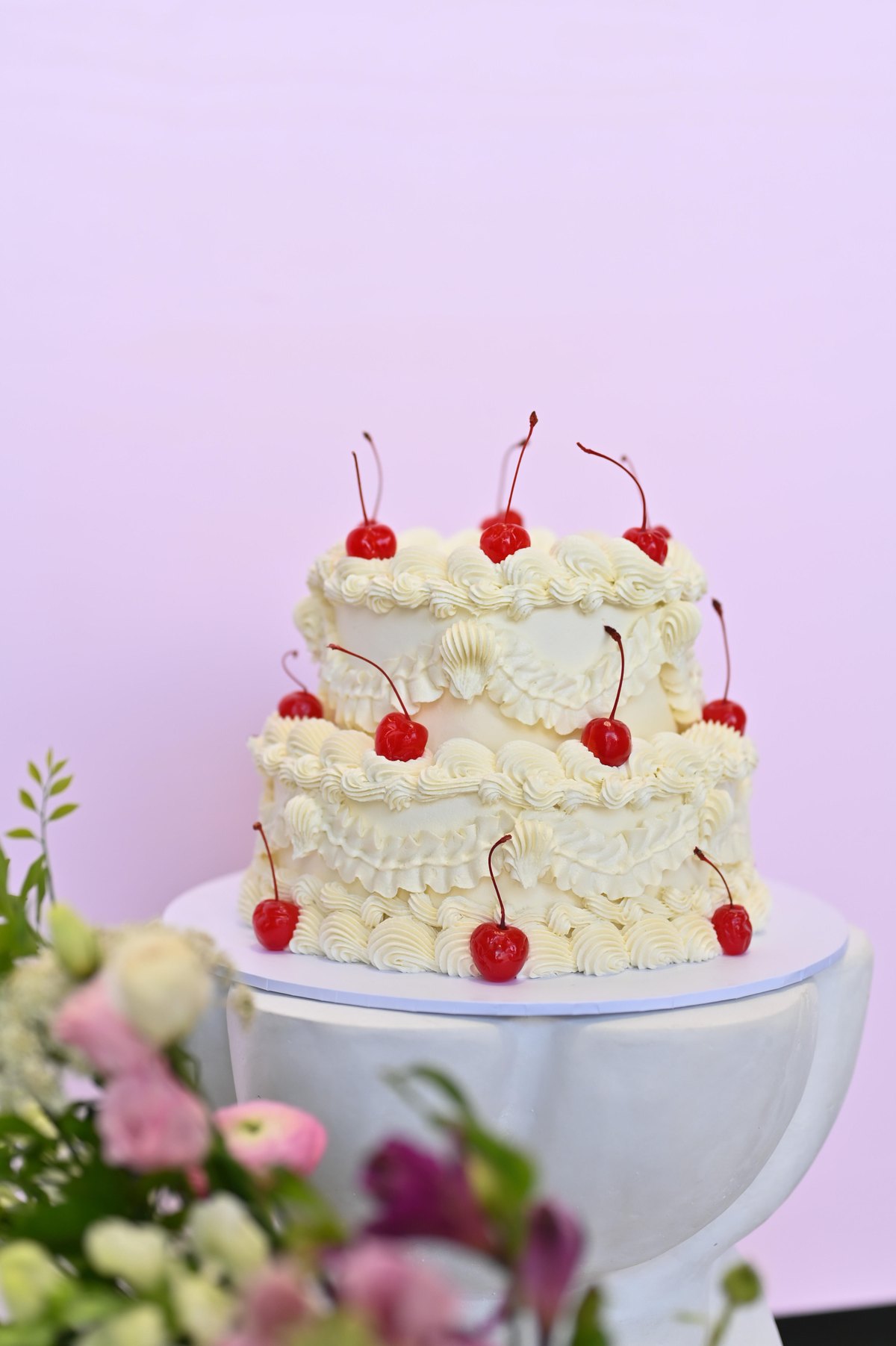 Two-tier vintage cream cake topped with maraschino cherries beside flowers